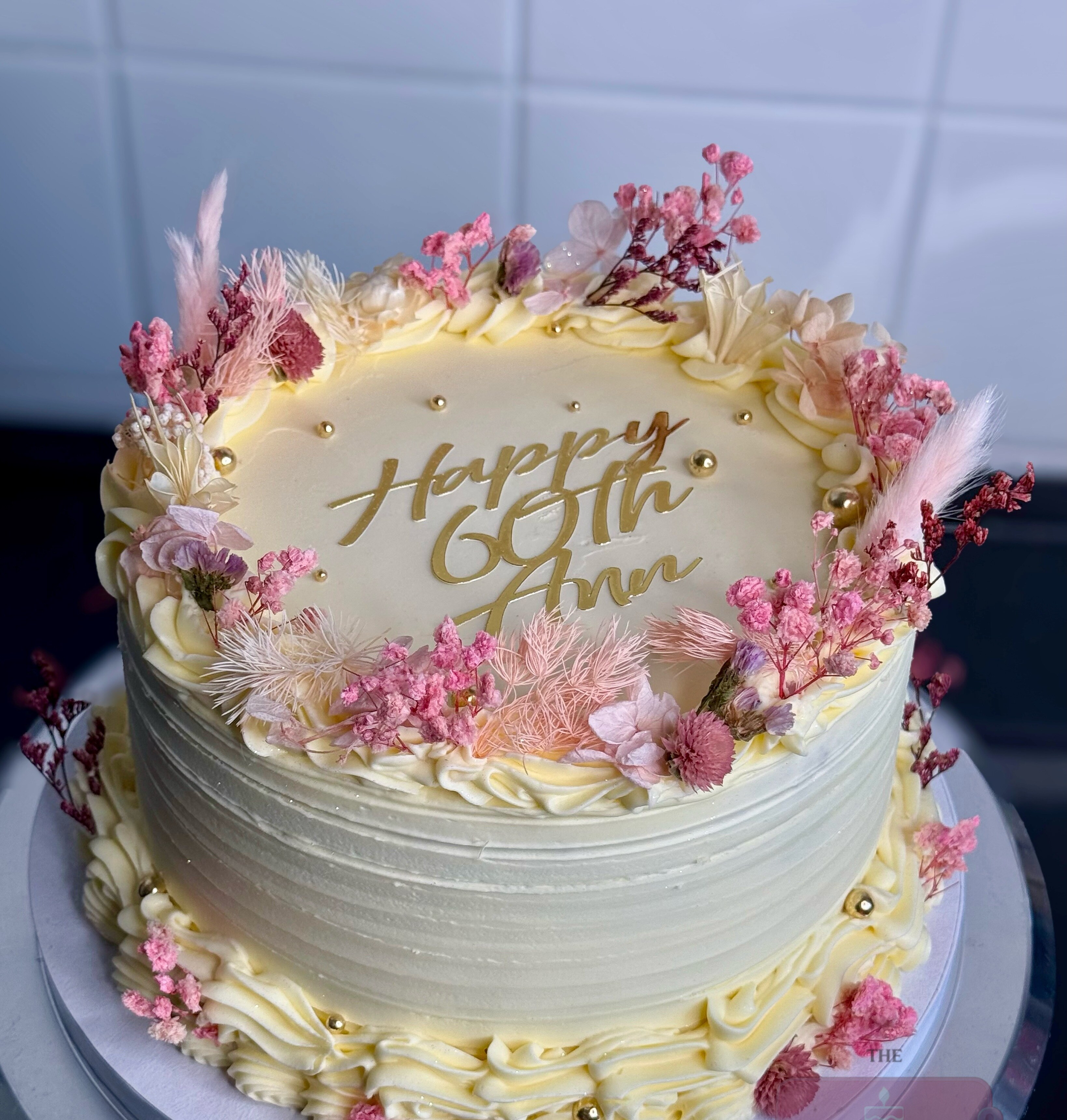 White celebration cake edged with dried pink flowers and gold lettering